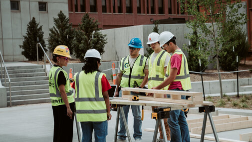 Construction workers in safety vests and helmets gather around a wood project outdoors.