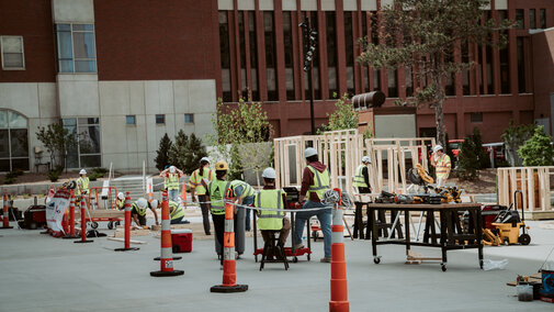 Construction workers in safety vests building wooden structures outdoors on a sunny day.